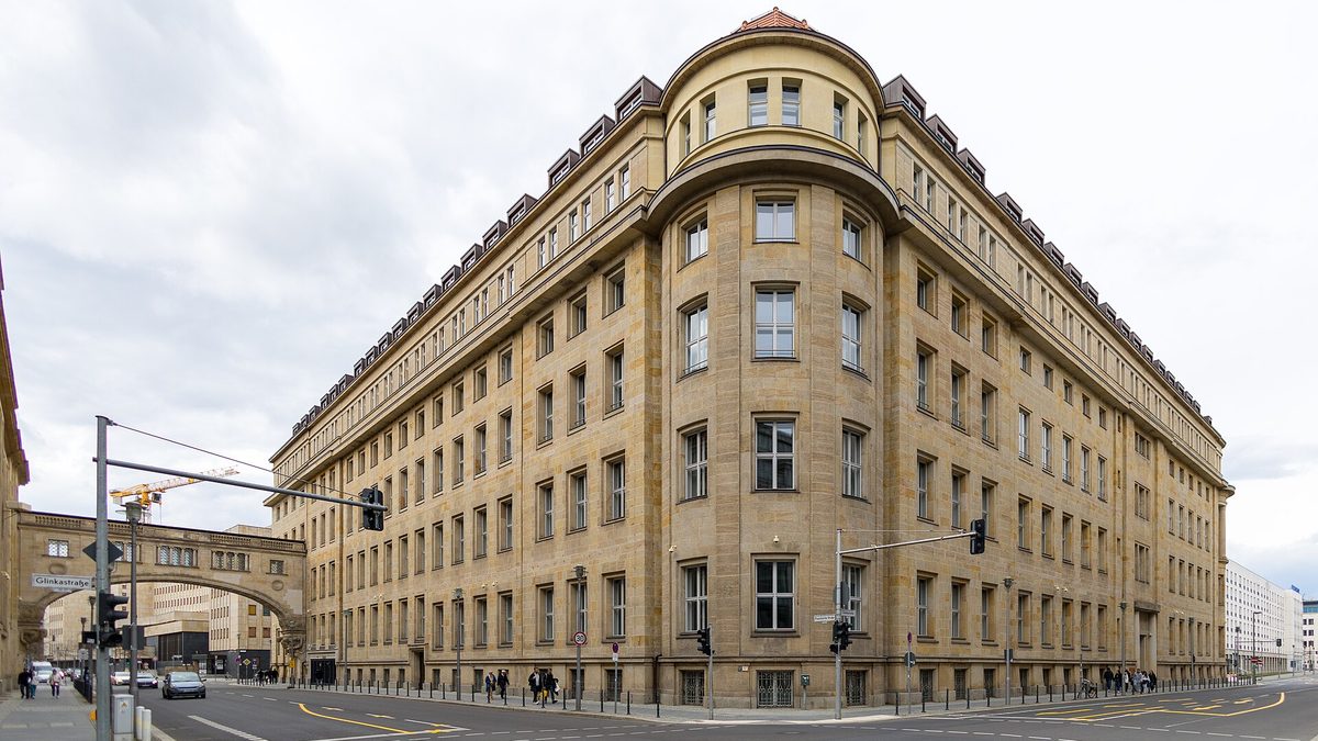 Ein großes Eckgebäude aus hellem Sandstein mit abgerundetem Eckturm, langen Fensterreihen und Dachgauben an einer Straßenkreuzung mit Ampeln; links eine steinerne Brücke zum Nachbargebäude, darüber bewölkter Himmel.