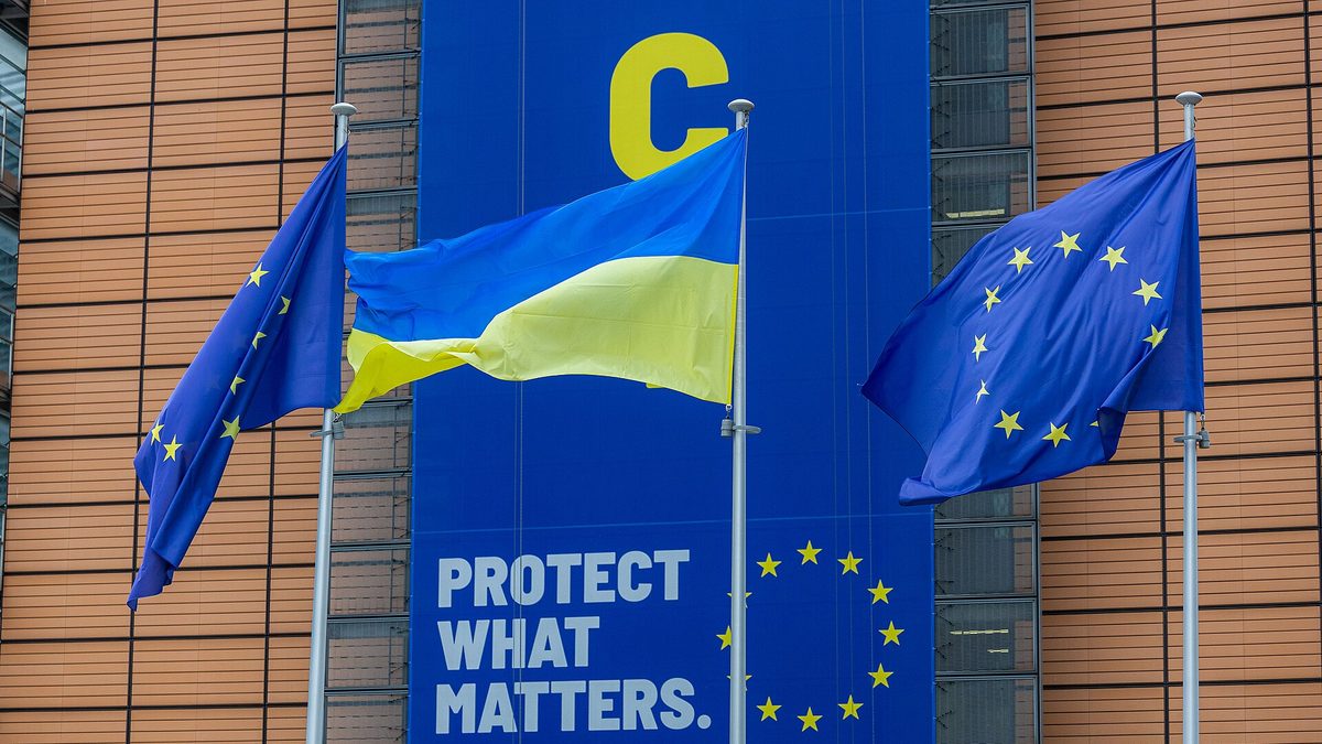 Die Flagge der Ukraine zwischen zwei EU-Flaggen an Fahnenmasten vor dem Berlaymont-Gebäude in Brüssel; dahinter ein großes blaues Transparent mit dem Schriftzug „Protect what matters.“ und dem Sternenkranz der EU.