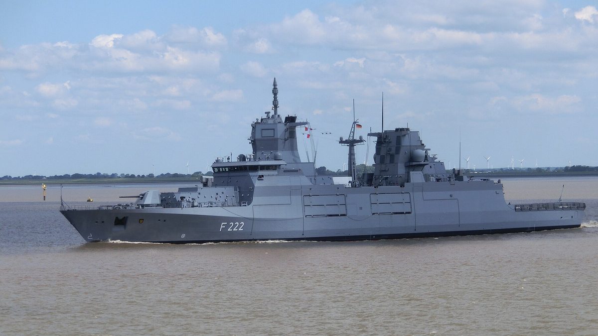 Ein graues Kriegsschiff mit der Kennung F 222 fährt auf bräunlichem Wasser nach links; am flachen Ufer im Hintergrund stehen Windräder, darüber blauer Himmel mit Quellwolken.