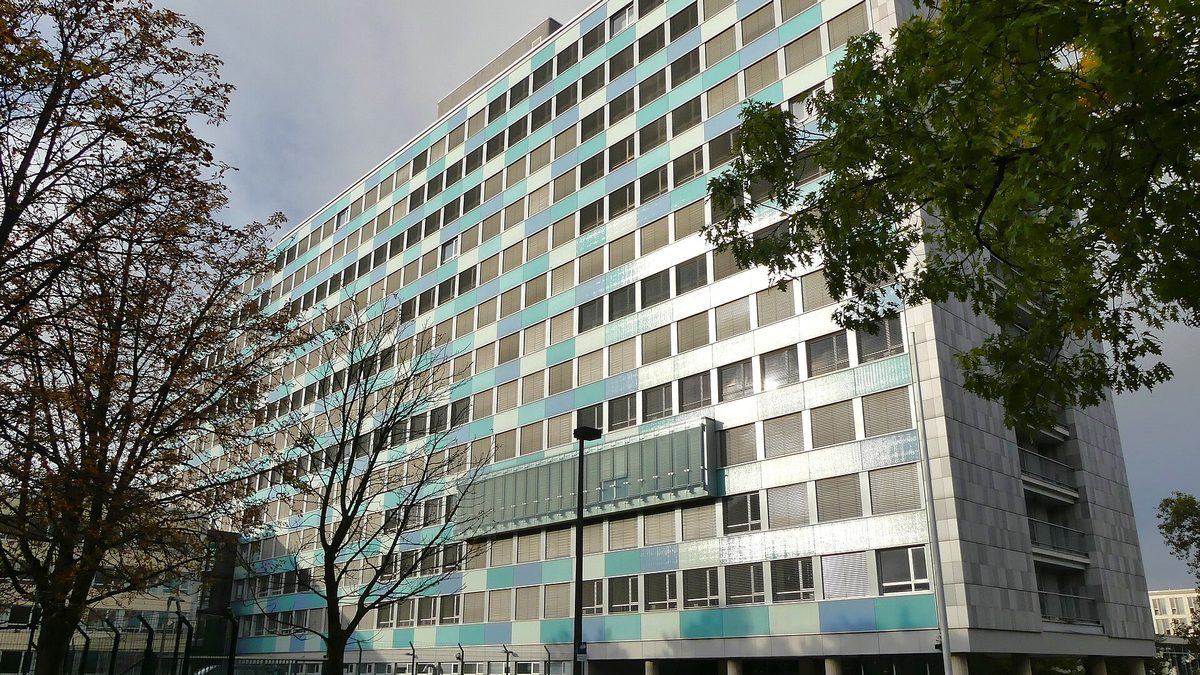 The high-rise of the Federal Statistical Office in Wiesbaden, its facade clad in white, pale green and blue panels between long rows of windows; in front a street lamp and trees, brown or bare on the left and still green on the right, under a cloudy sky.