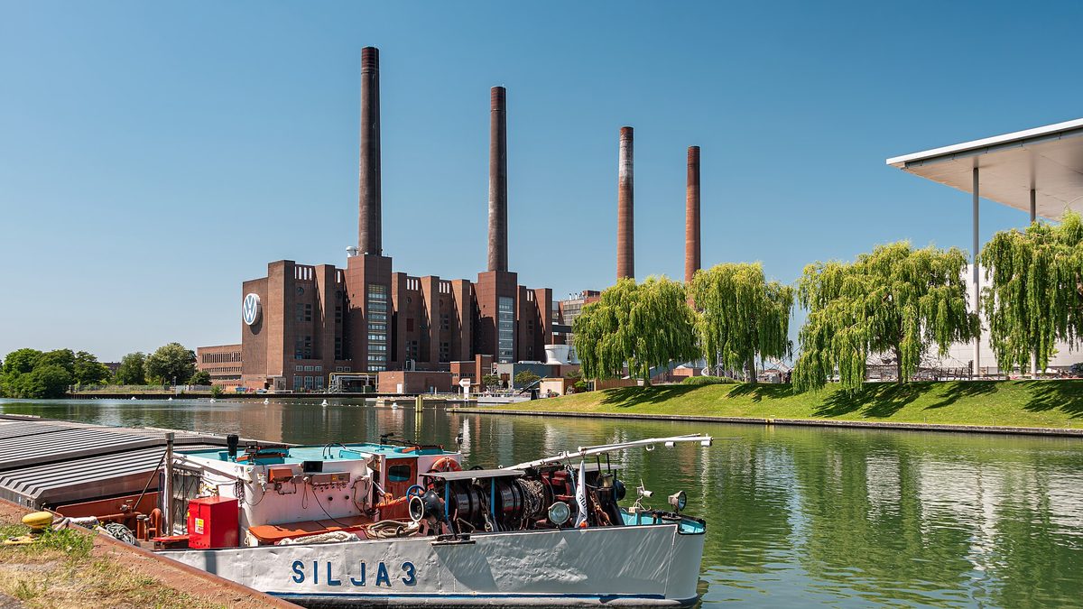 Das Heizkraftwerk des Volkswagenwerks in Wolfsburg am Mittellandkanal: ein Backsteinbau mit großem VW-Logo und vier hohen Schornsteinen unter blauem Himmel, rechts Trauerweiden am Ufer und im Vordergrund ein festgemachtes Binnenschiff mit dem Namen „Silja 3“.