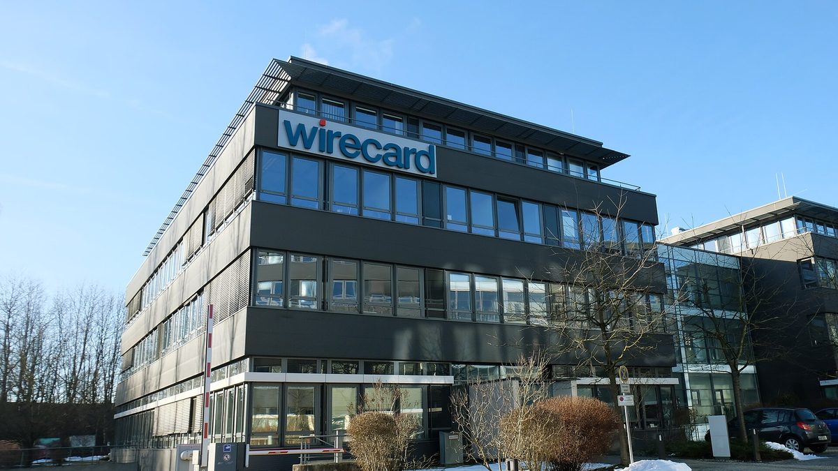 Ein dunkles, vierstöckiges Bürogebäude mit langen Fensterreihen und dem Schriftzug „wirecard“ am obersten Stockwerk, davor eine Schranke, kahle Sträucher und Schneereste, rechts ein Glasbau und parkende Autos, darüber blauer Himmel.