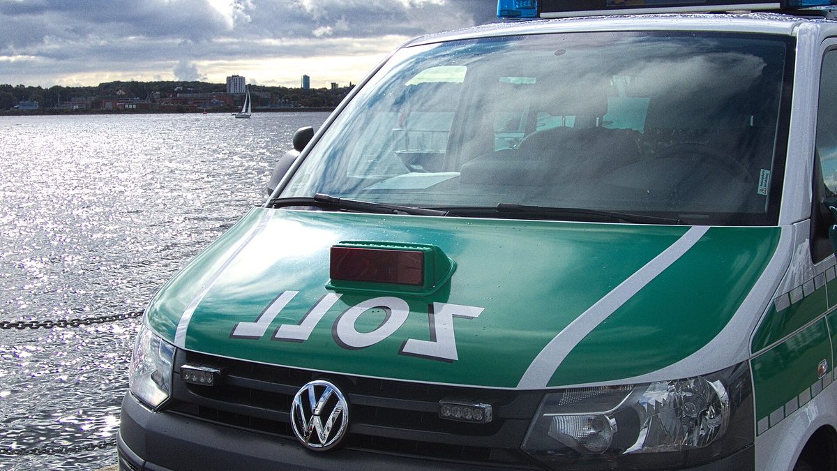 Die Front eines grün-weißen Kleinbusses des Zolls mit Blaulicht auf dem Dach und dem spiegelverkehrt aufgedruckten Schriftzug „ZOLL“ auf der Motorhaube, abgestellt an einer Kaimauer; dahinter Wasser, ein Segelboot und das gegenüberliegende Ufer unter Wolken.